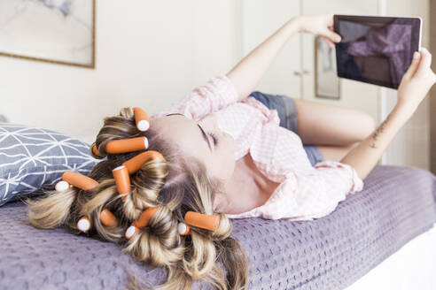 Young woman with curlers in hair lying on bed taking selfie with digital tablet - SDAHF00439