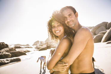 Glückliches Paar, das sich am Strand umarmt, mit Kindern im Hintergrund - SDAHF00465