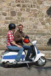 Lachendes Paar auf Motorroller vor einer Steinmauer, Italien - GIOF07986