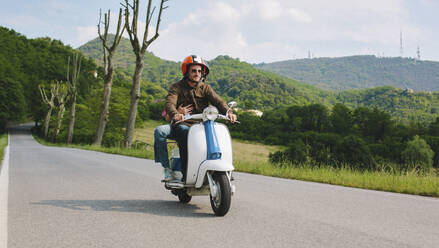 Couple riding vintage motor scooter on country road, Tuscany, Italy - GIOF07988