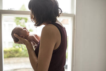 Mother cradling newborn baby son at window - HOXF04926