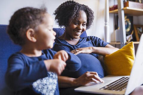 Glückliche schwangere Frau mit Tochter, die einen Laptop auf dem Sofa benutzt, lizenzfreies Stockfoto