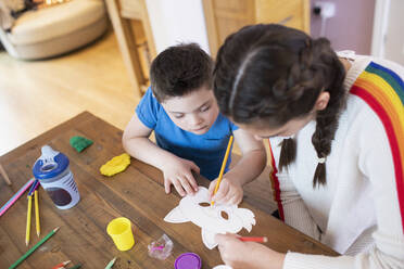 Girl helping brother with Down Syndrome color at table - HOXF04977