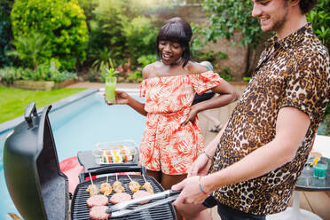 Young multiethnic couple barbecuing at poolside - HOXF05029