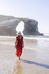 Blonde Frau in rotem Kleid und Hut, die am Strand spazieren geht, Natural Arch am Playa de Las Catedrales, Spanien - DGOF00380