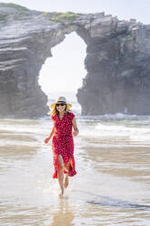 Blonde Frau in rotem Kleid und Hut, die am Strand läuft, Natural Arch am Playa de Las Catedrales, Spanien - DGOF00381