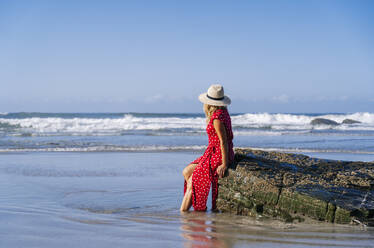 Blond woman wearing red dress and hat sittig on rock at the beach, Playa de Las Catedrales, Spain - DGOF00383