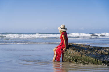 Blonde Frau in rotem Kleid und Hut auf einem Felsen am Strand sitzend, Playa de Las Catedrales, Spanien - DGOF00383