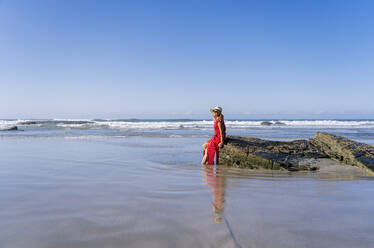 Blond woman wearing red dress and hat sittig on rock at the beach, Playa de Las Catedrales, Spain - DGOF00384