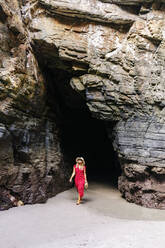 Blonde Frau in rotem Kleid und Hut in einer Felshöhle, Playa de Las Catedrales, Spanien - DGOF00389