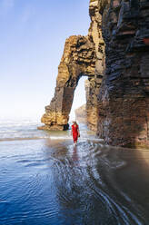 Blonde Frau in rotem Kleid und Hut, die am Strand spazieren geht, Natural Arch am Playa de Las Catedrales, Spanien - DGOF00391