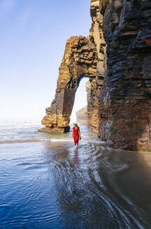 Blond woman wearing red dress and hat and walking along beach, Natural Arch at Playa de Las Catedrales, Spain - DGOF00391