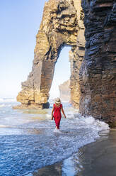 Blond woman wearing red dress and hat and walking along beach, Natural Arch at Playa de Las Catedrales, Spain - DGOF00392