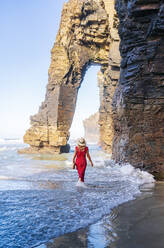 Blonde Frau in rotem Kleid und Hut, die am Strand spazieren geht, Natural Arch am Playa de Las Catedrales, Spanien - DGOF00392