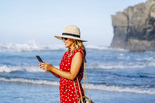 Blonde Frau in rotem Kleid und Hut, die am Strand ihr Smartphone benutzt - DGOF00395