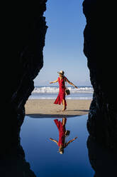 Blonde Frau in rotem Kleid und Hut, die am Strand tanzt, Playa de Las Catedrales, Spanien - DGOF00401