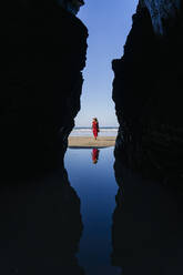Blonde Frau in rotem Kleid und Hut, die am Strand spazieren geht, Natural Arch am Playa de Las Catedrales, Spanien - DGOF00402