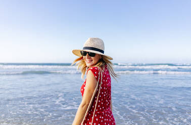 Blond woman wearing red dress and hat at the beach, turning and looking at camera - DGOF00407