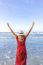 Blond woman wearing red dress and hat and walking along beach, Playa de Las Catedrales, Spain - DGOF00408