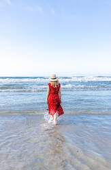 Rückansicht einer blonden Frau mit rotem Kleid und Hut, die am Strand ins Wasser geht - DGOF00409