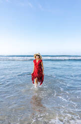 Blonde Frau in rotem Kleid und Hut am Strand, die im Wasser läuft - DGOF00410