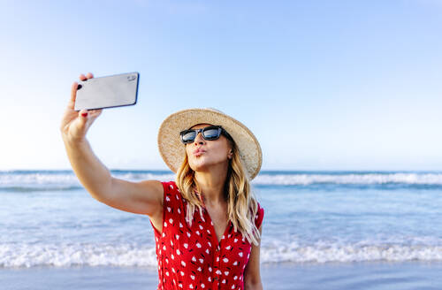 Blonde Frau in rotem Kleid und Hut, die ein Smartphone benutzt und ein Selfie am Strand macht, wobei sie einen Kussmund macht - DGOF00411