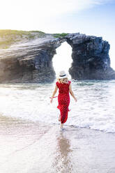 Blonde Frau in rotem Kleid und Hut, die am Strand spazieren geht, Natural Arch am Playa de Las Catedrales, Spanien - DGOF00413