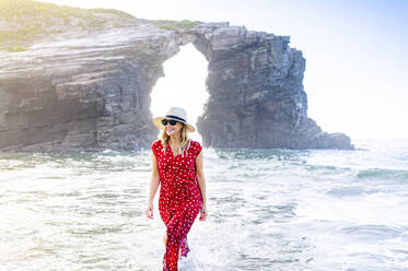 Blonde Frau in rotem Kleid und Hut, die am Strand spazieren geht, Natural Arch am Playa de Las Catedrales, Spanien - DGOF00414