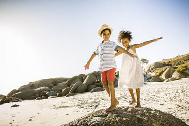 Happy boy and girl balancing on a rock on the beach - SDAHF00508