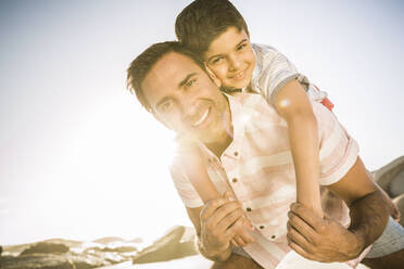 Portait of happy father carrying son piggyback on the beach - SDAHF00519