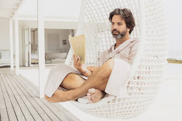 Relaxed man in bathrobe reading a book in hanging chair - SDAHF00531