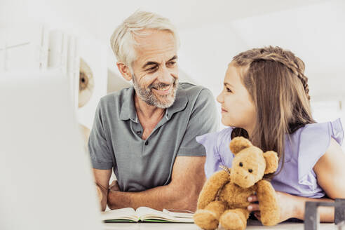 Grauhaariger Mann mit Laptop und Notebook, der ein Mädchen mit Teddybär zu Hause anlächelt - SDAHF00549