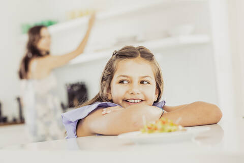 Porträt eines Mädchens, das sich an den Küchentisch lehnt, mit der Mutter im Hintergrund, lizenzfreies Stockfoto