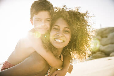 Portrait of a happy mother carrying her son piggyback on the beach - SDAHF00600