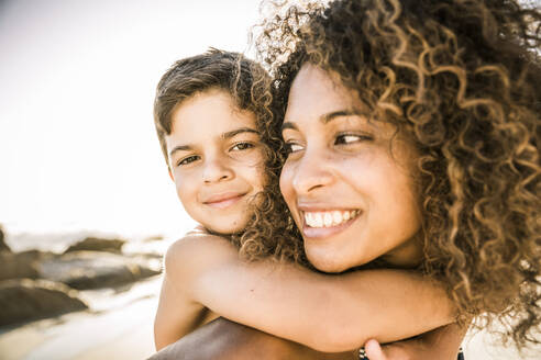 Portrait of a happy mother carrying her son piggyback on the beach - SDAHF00606