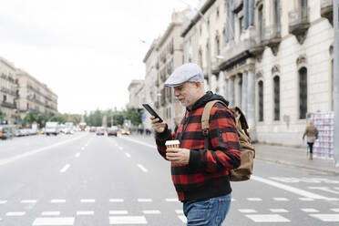 Älterer Mann mit Rucksack und Kaffee zum Mitnehmen schaut beim Überqueren der Straße auf sein Handy, Barcelona, Spanien - JCZF00009