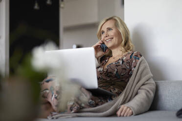 Blonde Frau mit Smartphone und Laptop auf der Couch zu Hause - RBF07059