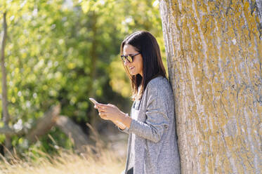 Lächelnde Frau mit Smartphone, an einen Baum in einem Park gelehnt - DGOF00433