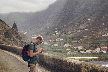 Spanien, La Gomera, Hermigua, Junger Mann überprüft Smartphone auf der Straße - MAMF01155