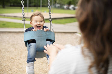 Mother pushing happy cute toddler girl in swing at playground - CAIF23792