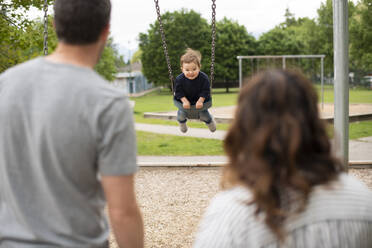 Eltern beobachten sorgloses Kleinkindmädchen beim Schaukeln auf dem Spielplatz - CAIF23794