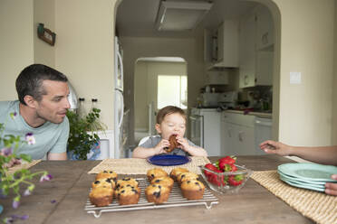 Father watching cute toddler daughter eating muffin at kitchen table - CAIF23808