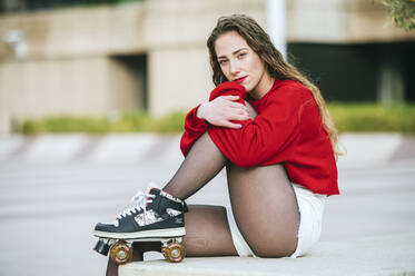 Porträt einer jungen Frau mit Rollschuhen, die auf einer Mauer in der Stadt sitzt - KIJF02909
