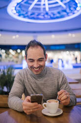 Man sitting in cafeteria, drinking coffee, using smartphone - GRCF00174