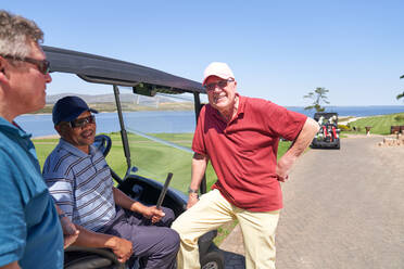 Glückliche männliche Golffreunde unterhalten sich am Golfwagen auf einem sonnigen Platz - CAIF23850