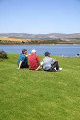Männliche Golfer machen eine Pause und ruhen sich im Gras auf einem sonnigen Golfplatz aus - CAIF23863