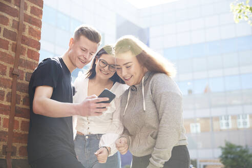 Freunde im Teenageralter benutzen ein Smartphone vor einem sonnigen Schulgebäude - CAIF24107
