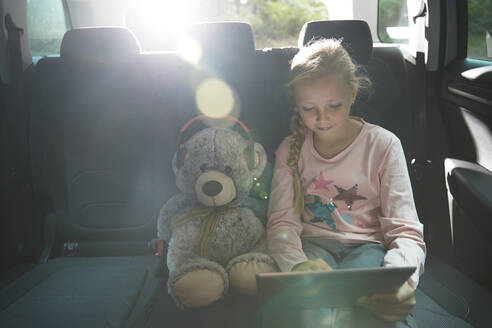 Mädchen mit Teddybär benutzt digitales Tablet auf dem Rücksitz eines Autos - CAIF24255