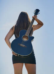 A teenage female guitarist holds an acoustic guitar to her back against a blue sky - CAVF74949