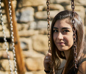 Portrait of happy teenage girl with brown hair is sitting on a swing - CAVF74954